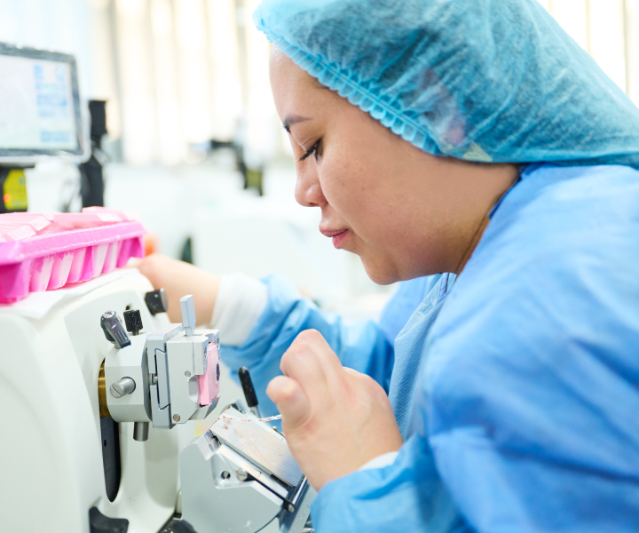 Una profesional de la salud vestida con bata azul y gorro, se encuentra sentada frente a un microscopio revisando muestras de laboratorio