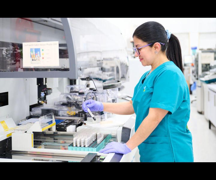 En la parte izquierda está un escritorio con tubos de ensayo, en la parte derecha una profesional de la salud tomando y revisando una  muestra en específico.